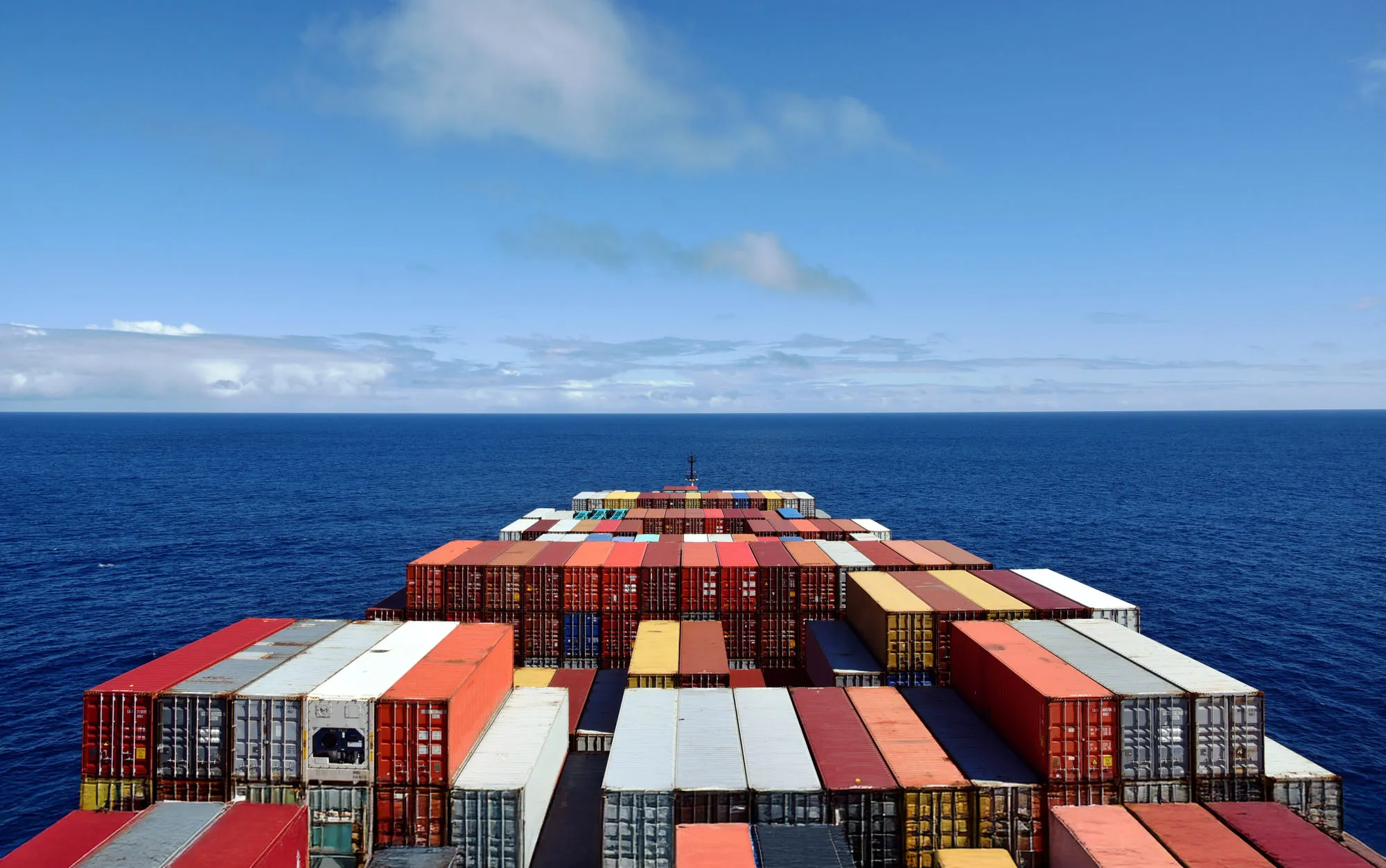 View from the stern of a cargo ship deck with colorful stacks of containers on the open sea under a blue sky.
