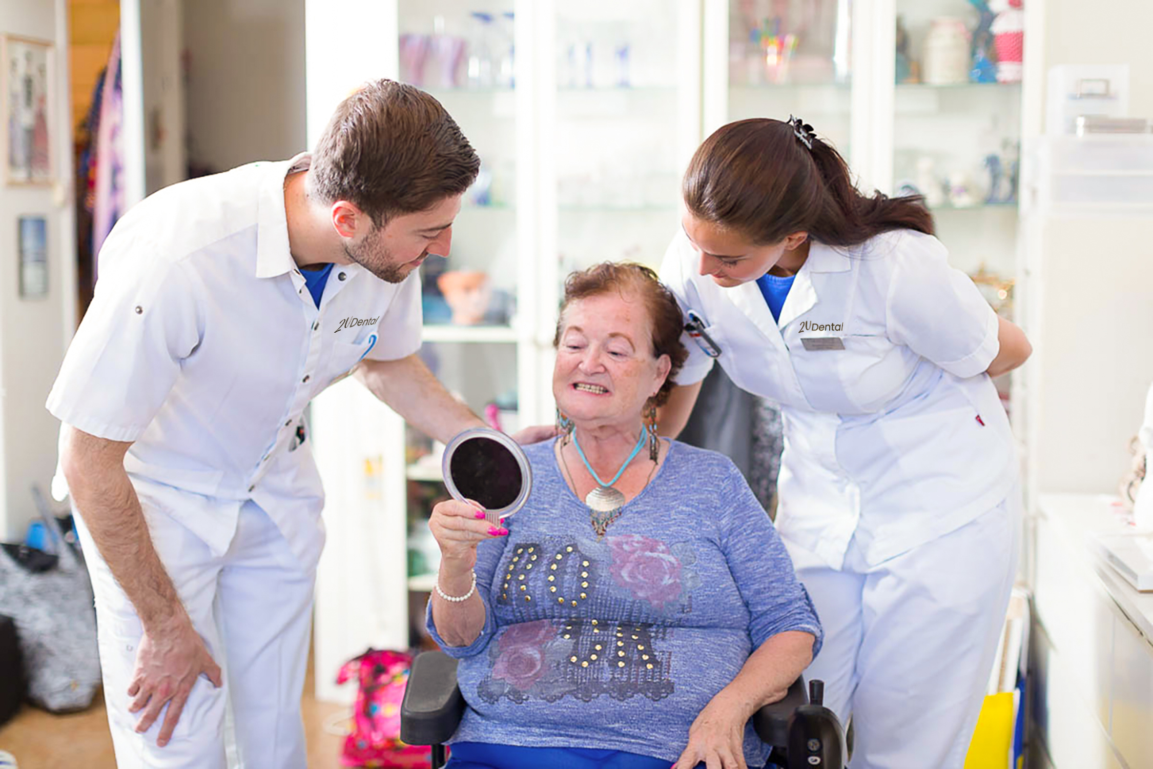 Two dental professionals wearing masks and gloves providing dental care to a smiling elderly woman seated in a recliner in a senior care facility.