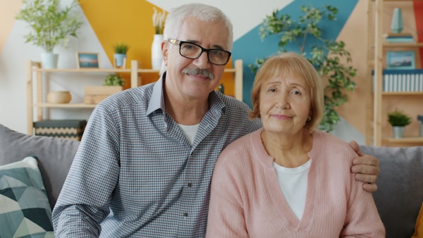 Elderly husband and wife are looking at camera with serious faces then smiling and laughing expressing positive emotions sitting on sofa at home.