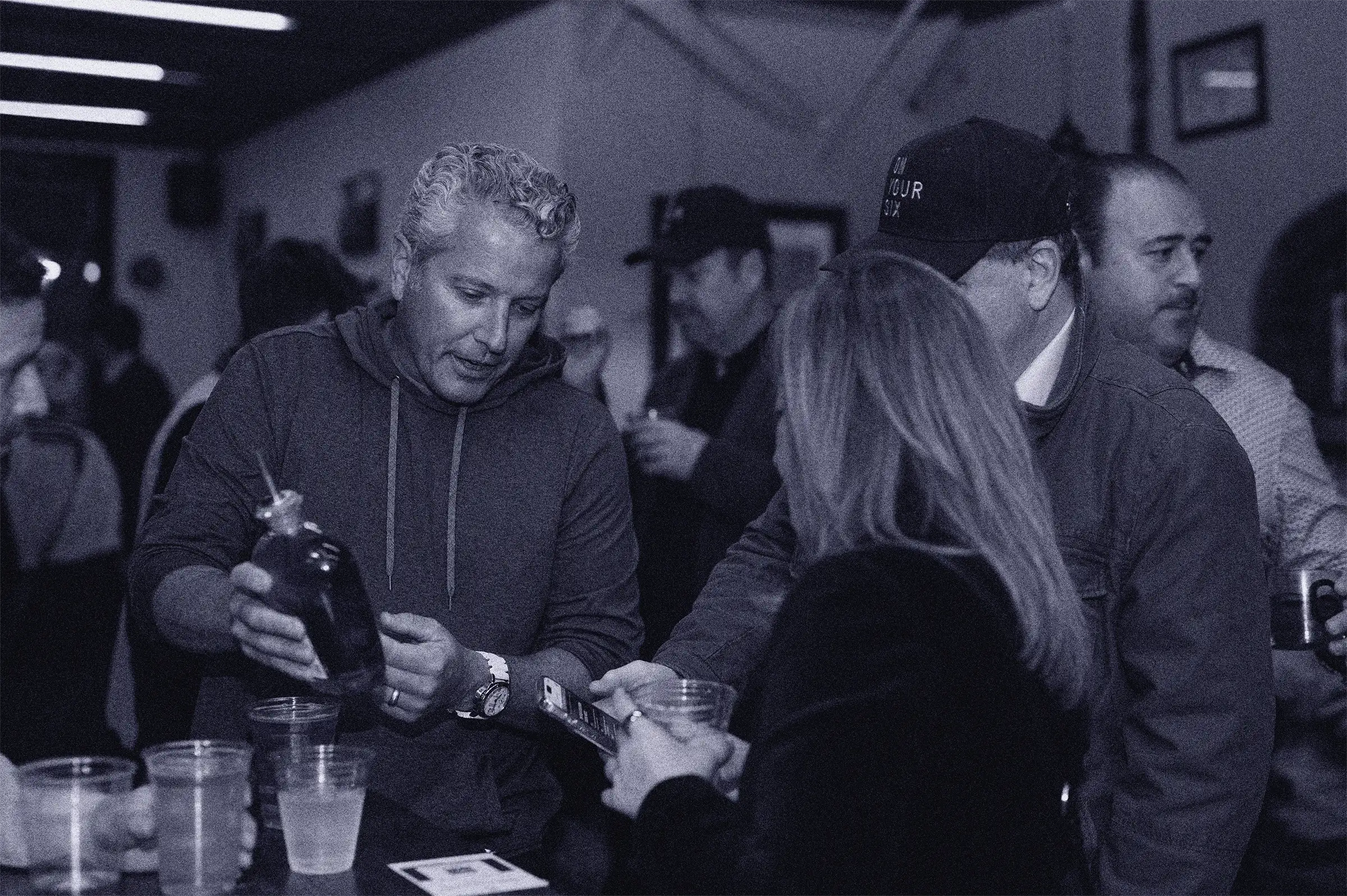 Group of people at a tasting event sampling On Your Six bourbon whiskey, with a man pouring a drink and others engaged with the brand experience.