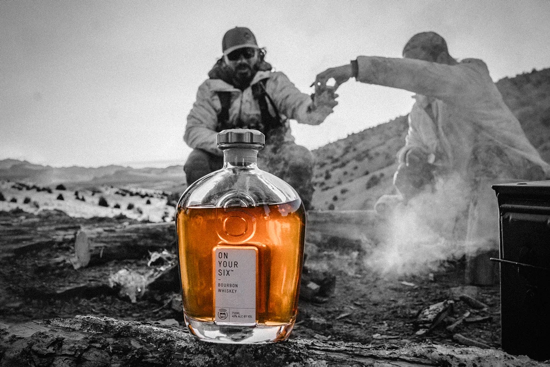 A bottle of On Your Six Bourbon sits on a log in front of three men.