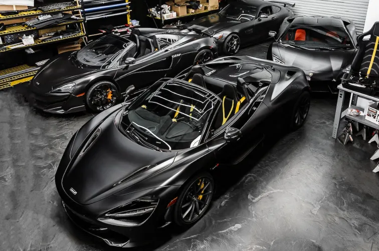 Four black luxury sports cars parked indoors on a polished concrete floor with shelves and automotive equipment in the background.