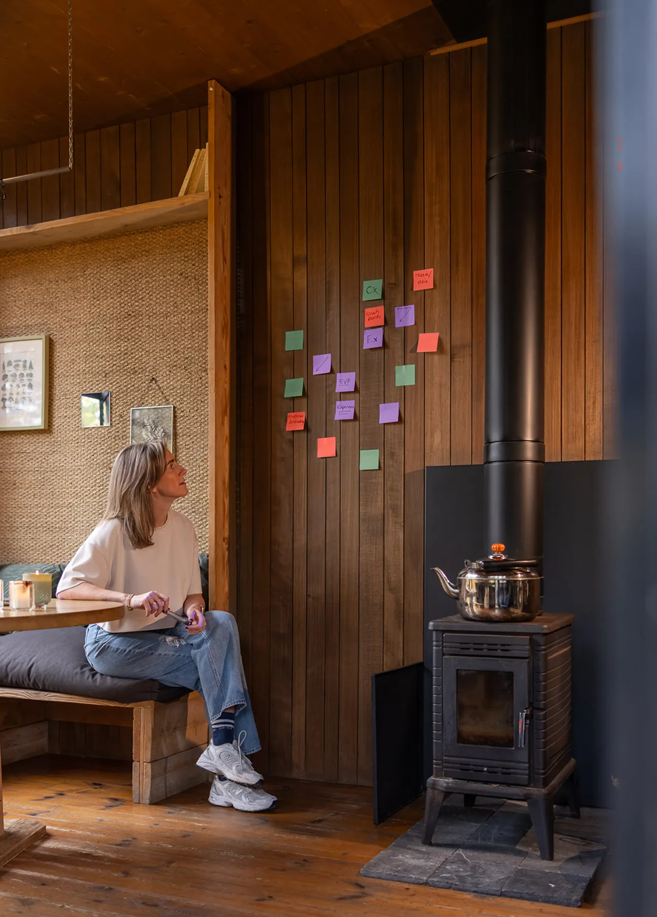 Woman sitting on a bench in a cozy wooden room looking at colorful sticky notes on a wooden wall beside a black wood stove with a kettle.