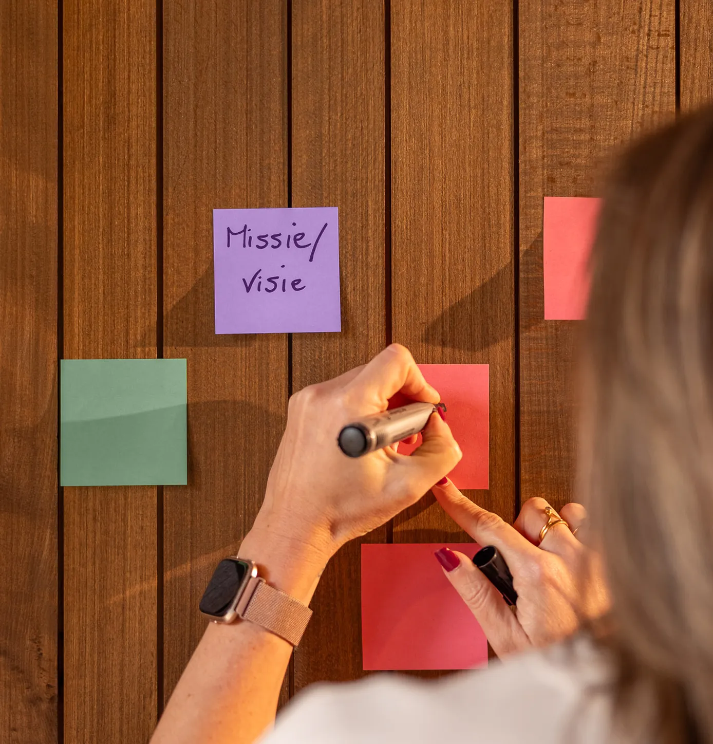 Person writing on a red sticky note on a wooden wall with other colorful sticky notes, including one labeled 'Missie/visie'.
