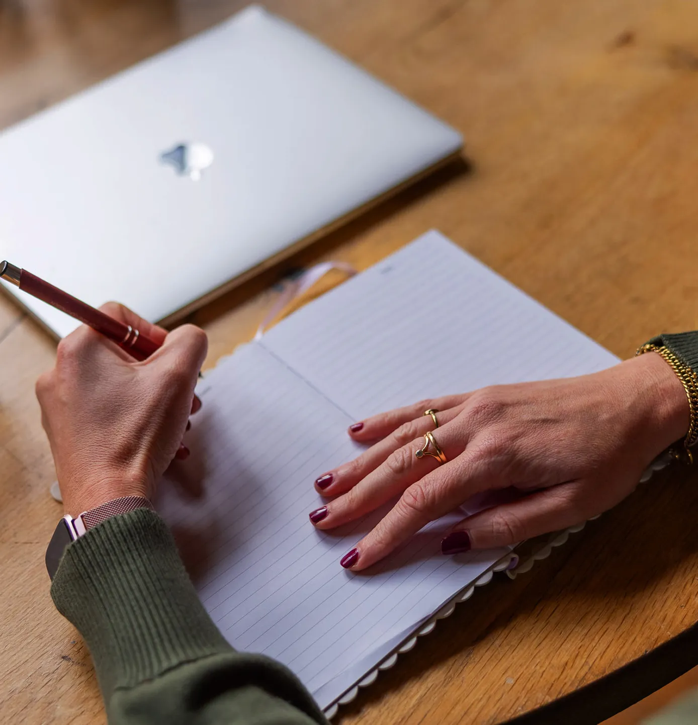 Person with maroon nail polish and gold rings writing in a lined notebook with a pen, a closed laptop on a wooden table.