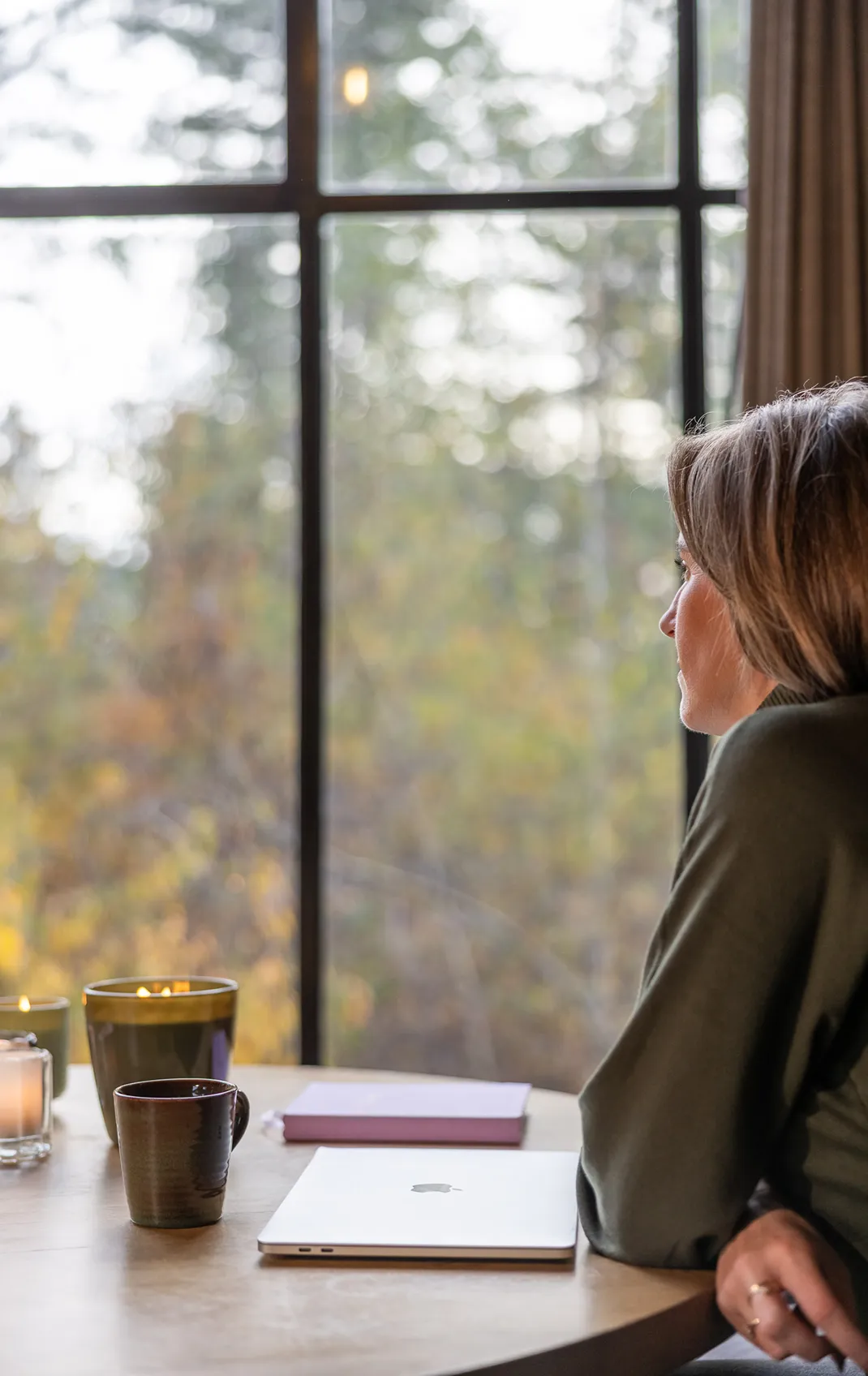 Woman in green sweater sitting at table with closed laptop, coffee mug, candles, and notebook, looking out a large window.