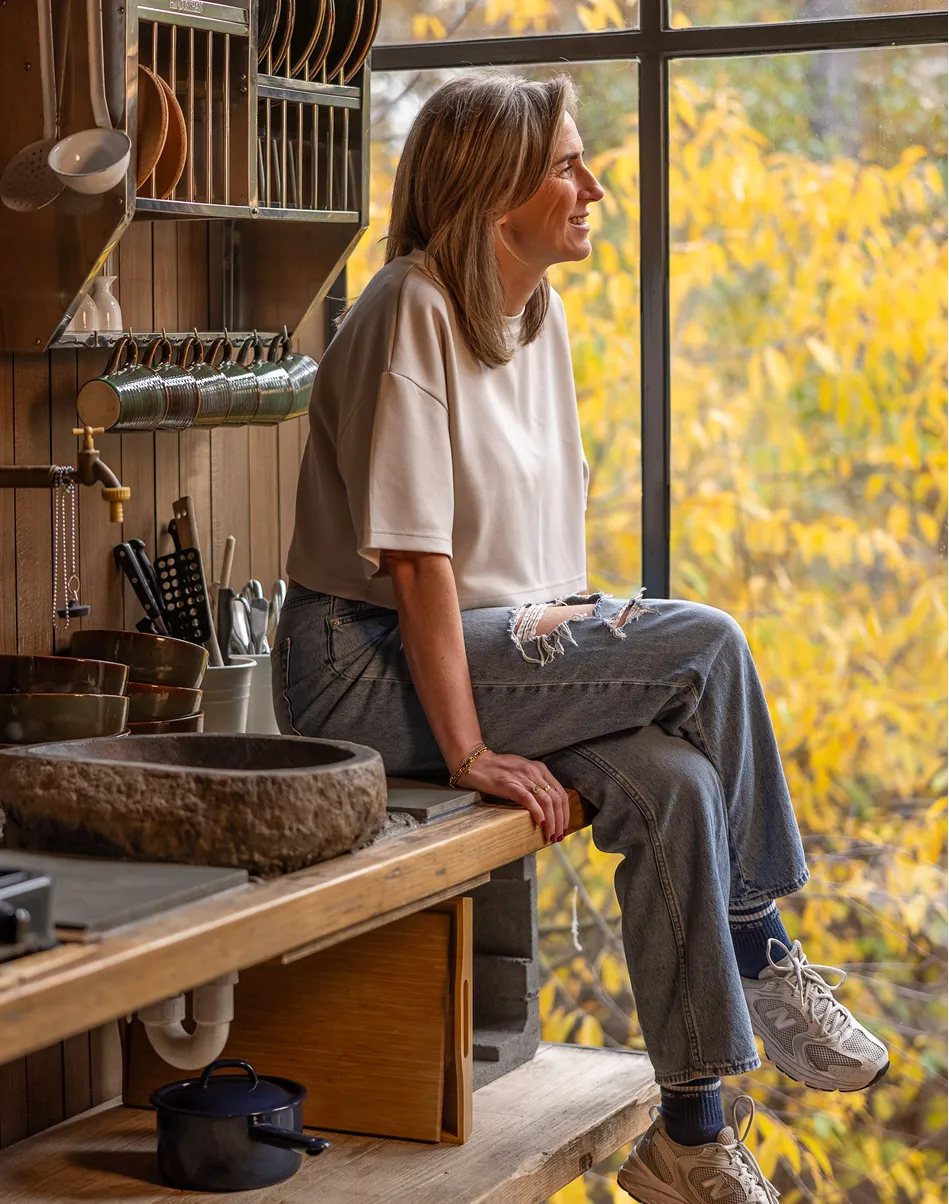 Woman in casual clothes sitting on a wooden kitchen counter, looking out a window with autumn foliage outside.
