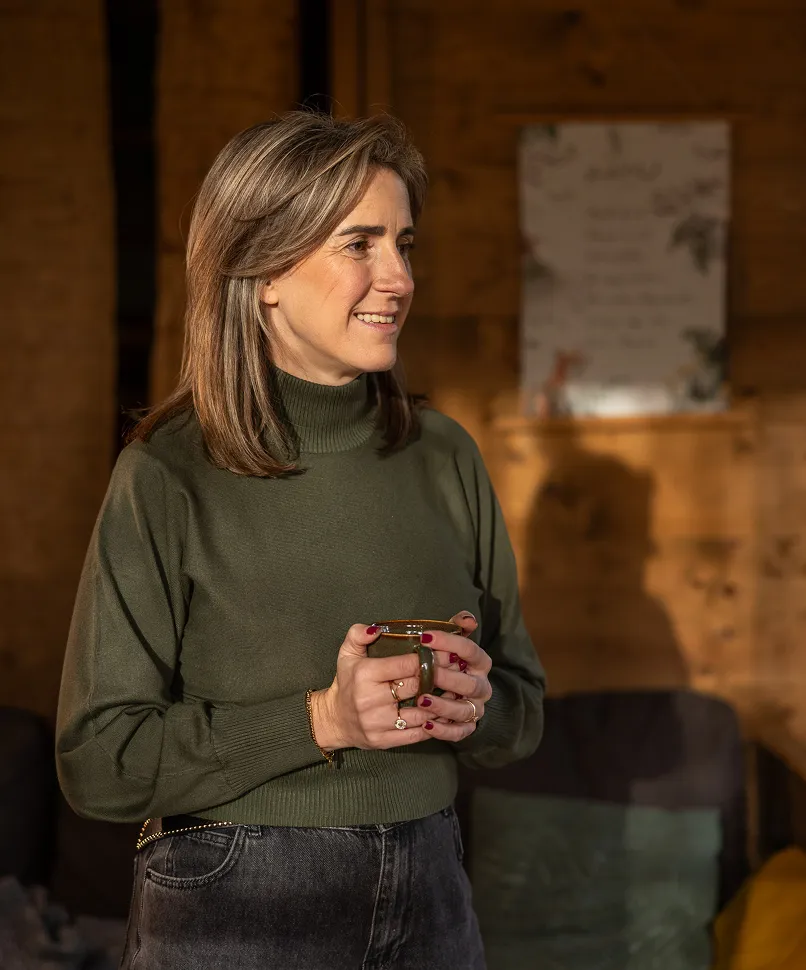 Woman in green turtleneck sweater holding a cup and smiling, standing indoors with wooden wall background.