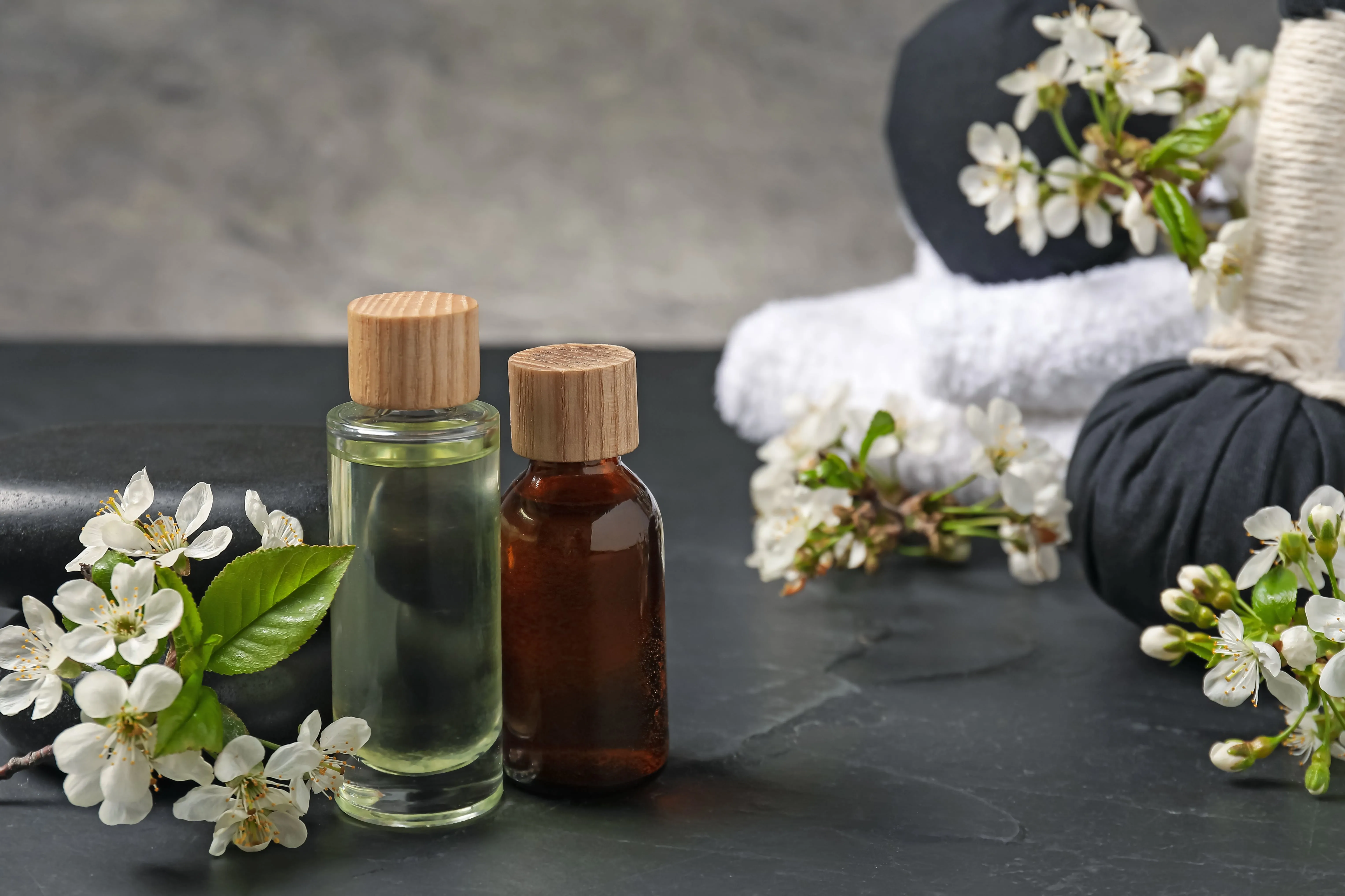 Zwei kleine Flaschen mit ätherischen Ölen neben weißen Blüten und einem schwarzen Stein auf dunklem Untergrund.