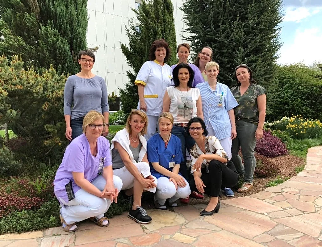 Gruppe von elf Frauen, darunter mehrere in medizinischer Berufsbekleidung, die in einem Garten mit Bäumen und Blumen posieren.