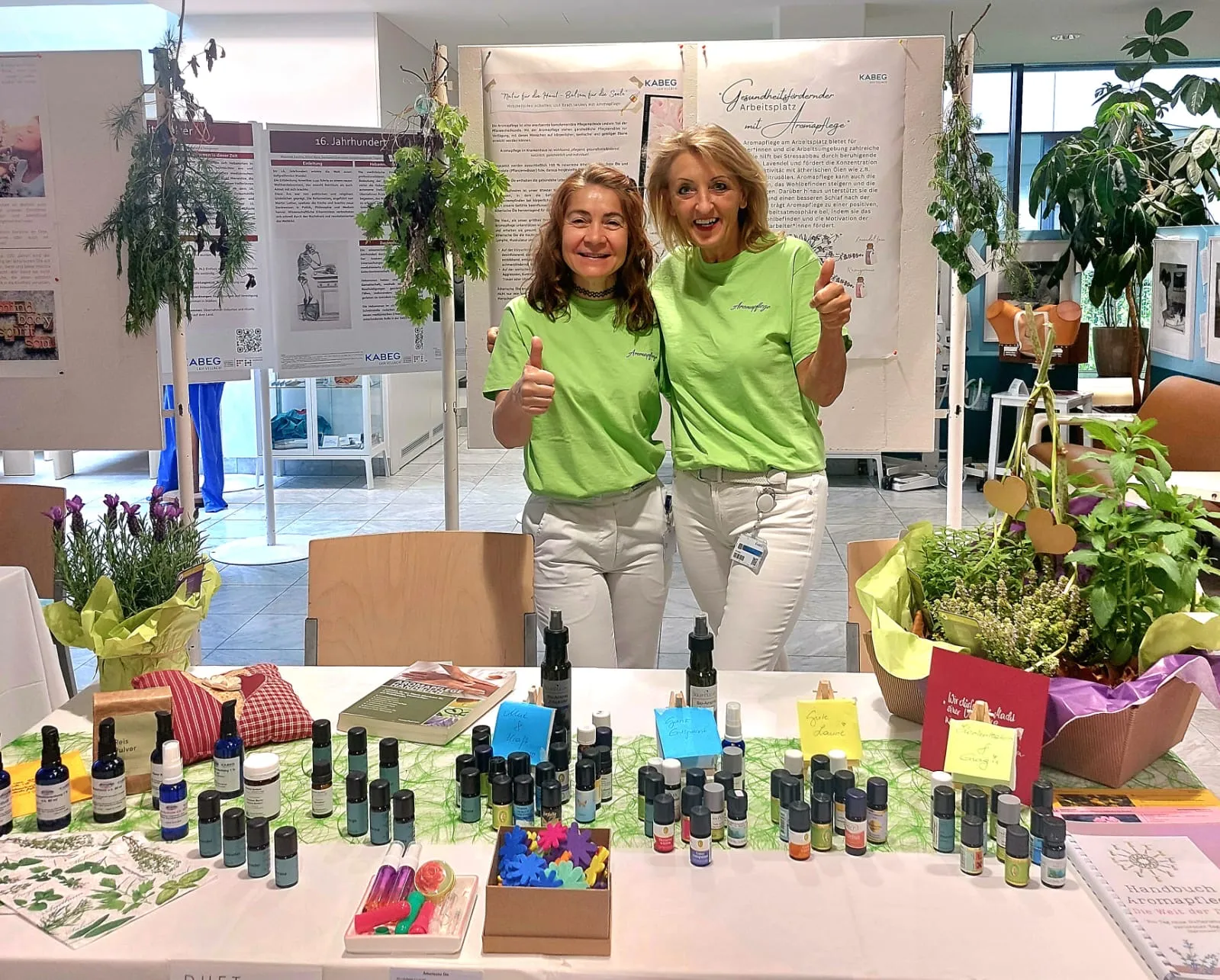 Lisa Walder und eine andere Frauen stehen in grünen T-Shirts hinter einem Tisch mit Aromapflege-Produkten und Pflanzen und zeigen den Daumen nach oben.