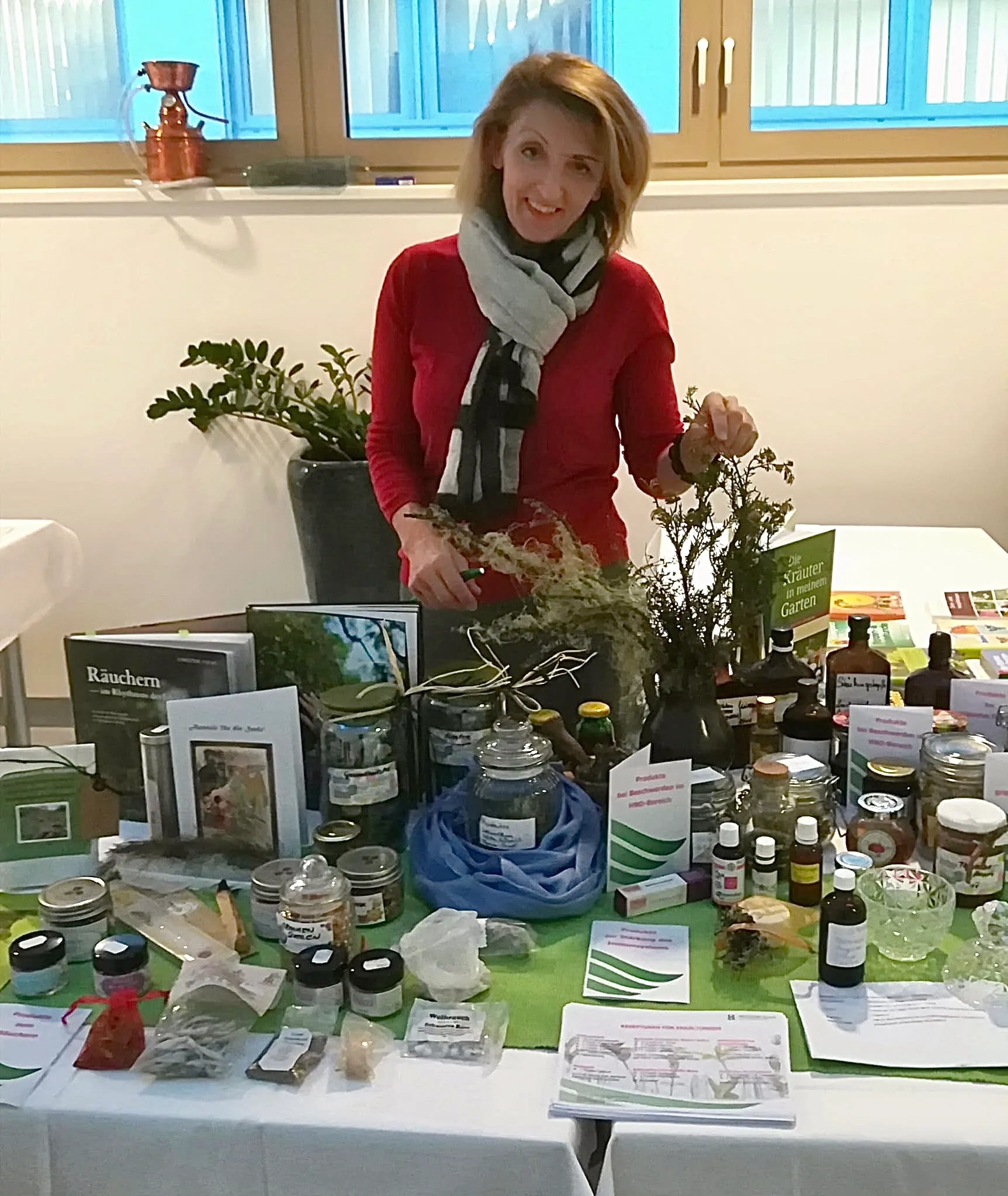 Lisa Walder steht hinter einem Tisch mit verschiedenen Kräuterprodukten, Büchern und Gläsern in einem hellen Raum.