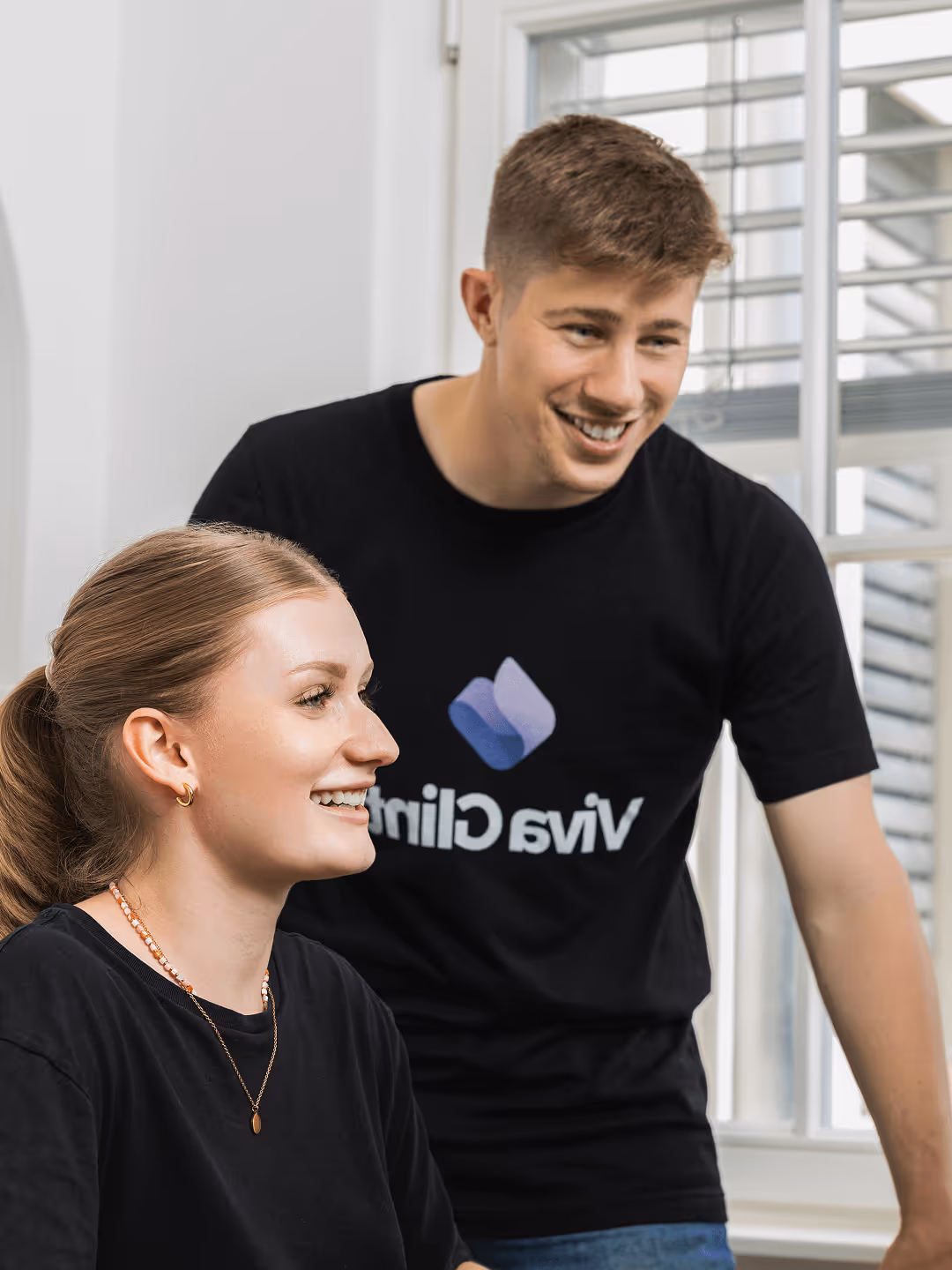 Zwei lächelnde Personen in schwarzen Viva Clinic T-Shirts in einem hellen Raum mit Fenster.