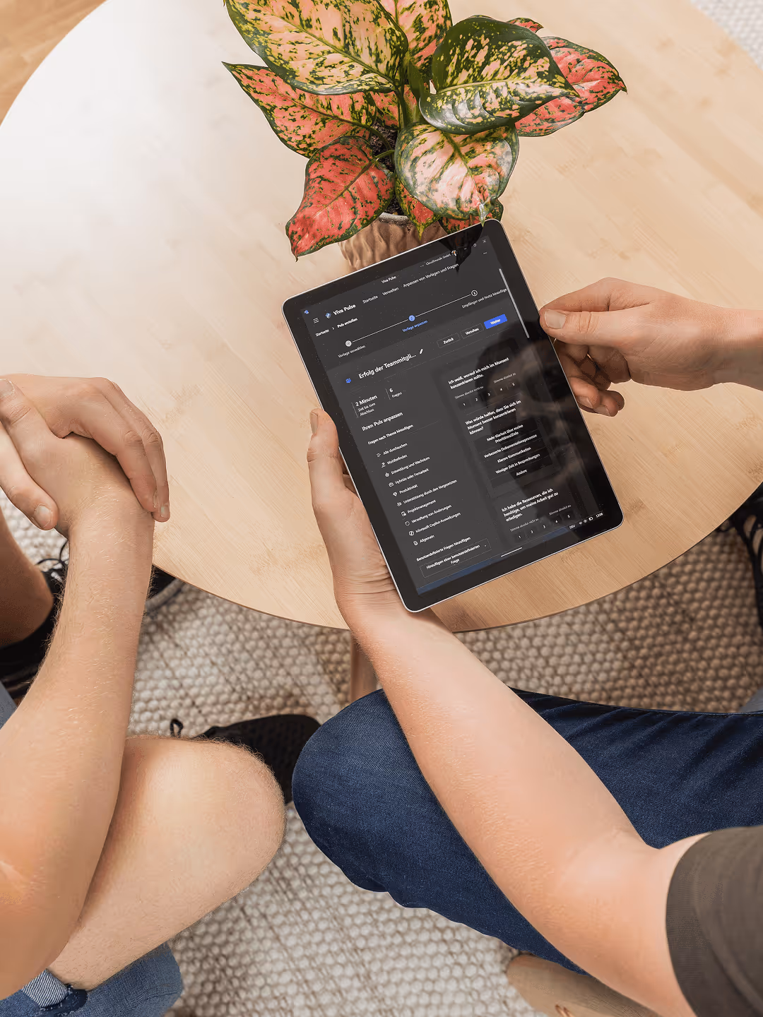 Zwei Personen sitzen an einem Tisch mit einer Pflanze, während eine Person ein Tablet mit einer Webseite nutzt.