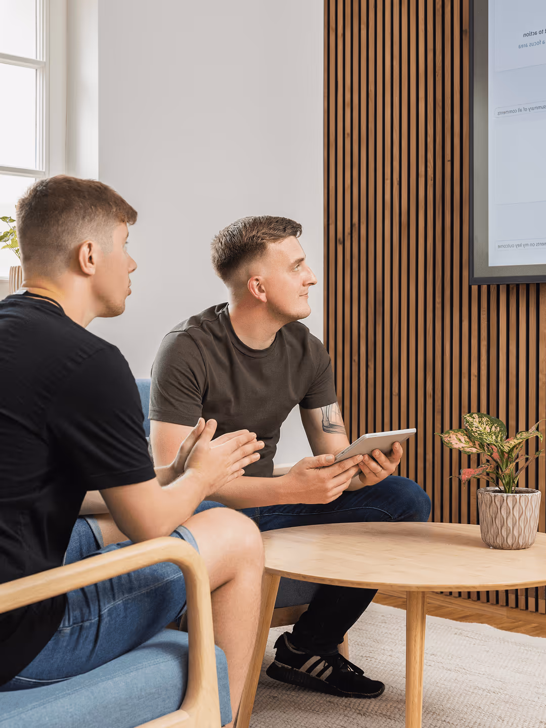 Zwei Männer in Freizeitkleidung sitzen in modernem Büro, diskutieren und betrachten Bildschirm, einer hält Tablet.