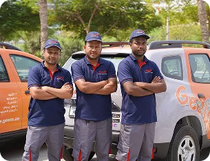 Three men in matching navy blue shirts and gray pants standing with arms crossed in front of vehicles with orange and white branding.