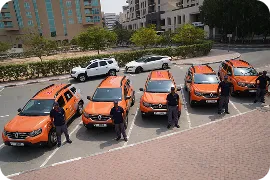 Five orange and black cars parked in a row with uniformed security personnel standing beside each car in an urban parking area.