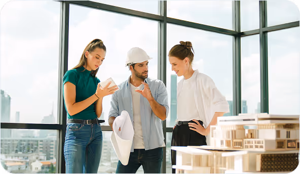Three architects discussing a building model in a modern glass office, one wearing a white hard hat and holding blueprints.