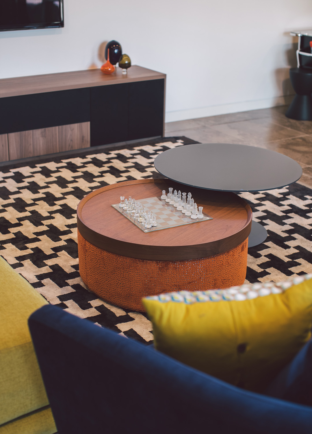 Modern living room with patterned rug, round wooden coffee table with glass chess set, and colorful cushions on sofa.