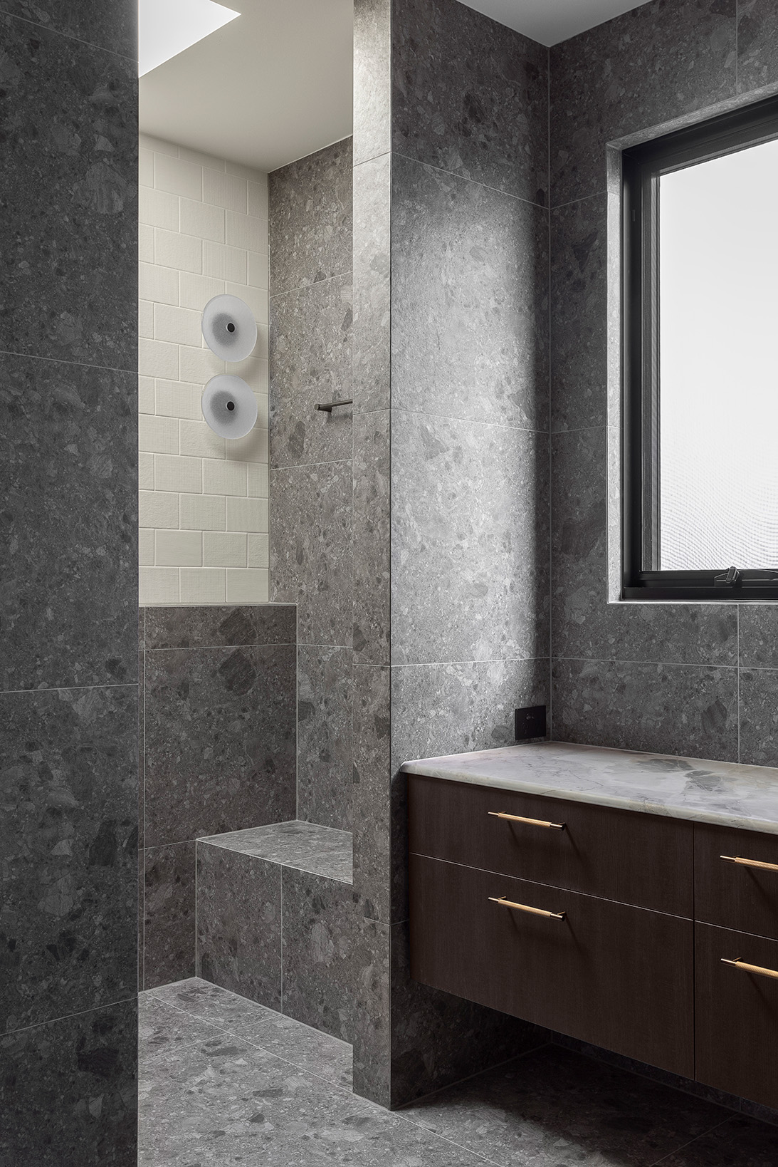 Modern bathroom corner with grey stone tiles, built-in shower bench, dark wood vanity with marble countertop, and a frosted window.