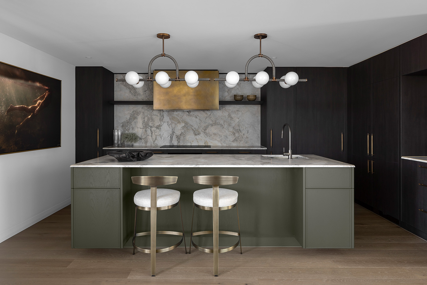 Modern kitchen with a marble countertop island, two brass bar stools, dark cabinetry, brass range hood, and a large underwater photo on the wall.