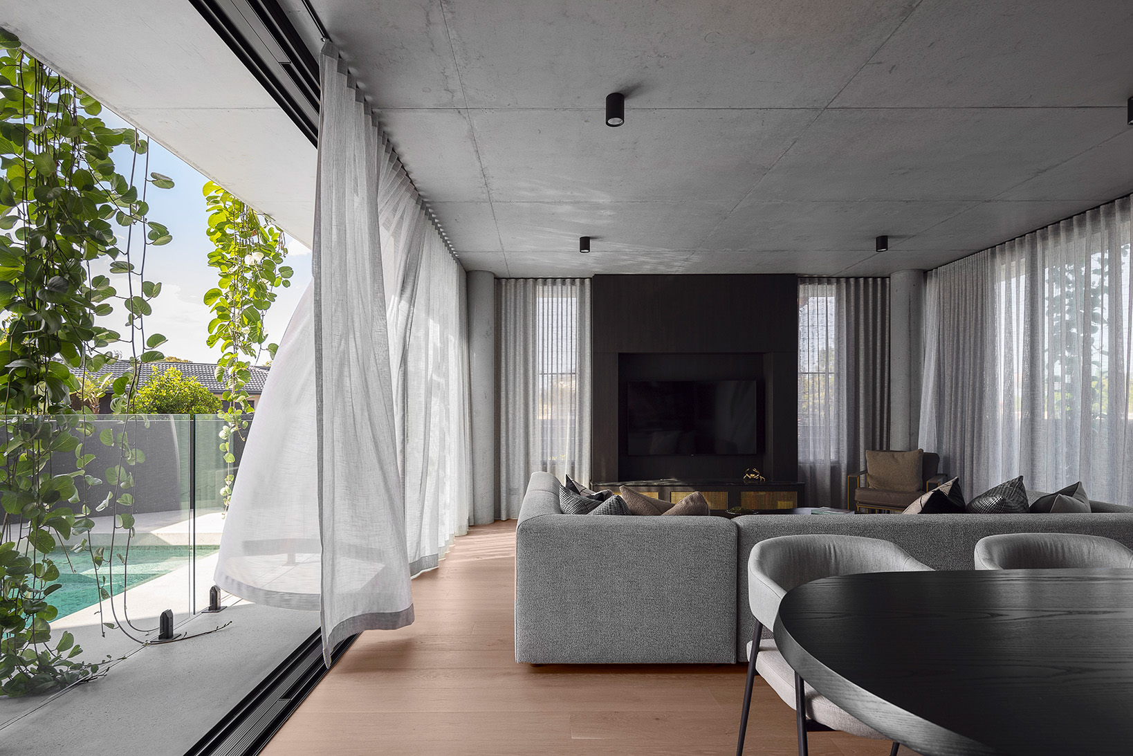 Modern living room with large gray sectional sofa, sheer curtains, a dark TV wall unit, and a view of a pool and greenery outside through floor-to-ceiling windows.