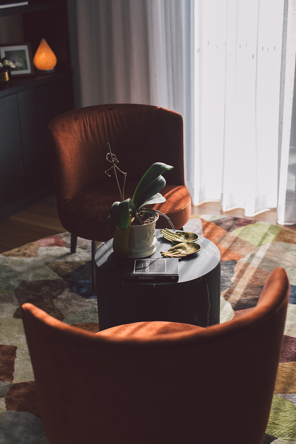 Cozy corner with two rust-colored velvet chairs around a small black round table holding a potted plant, a Chanel book, and two gold hand-shaped decor pieces, with sunlight streaming through white curtains.