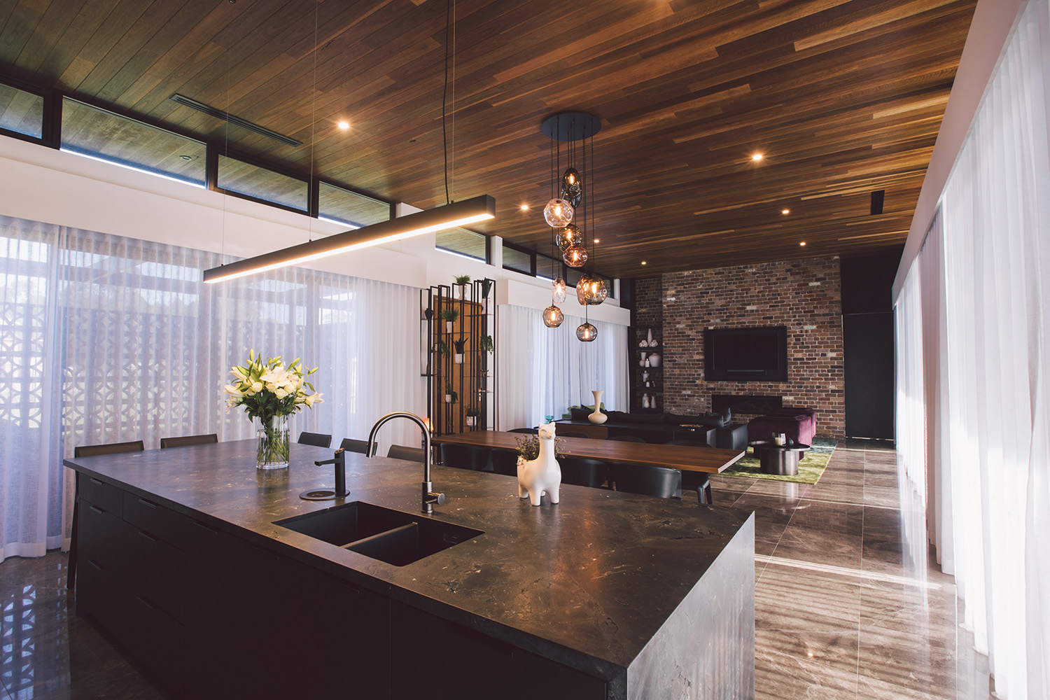 Modern open kitchen and living area with dark stone island, wooden ceiling, hanging pendant lights, and brick accent wall with TV.