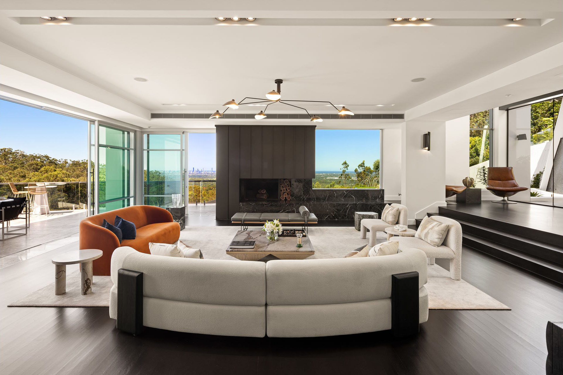 Modern living room with curved white sofa, orange loveseat, white chairs, black fireplace, and large windows showing green trees and cityscape beyond.