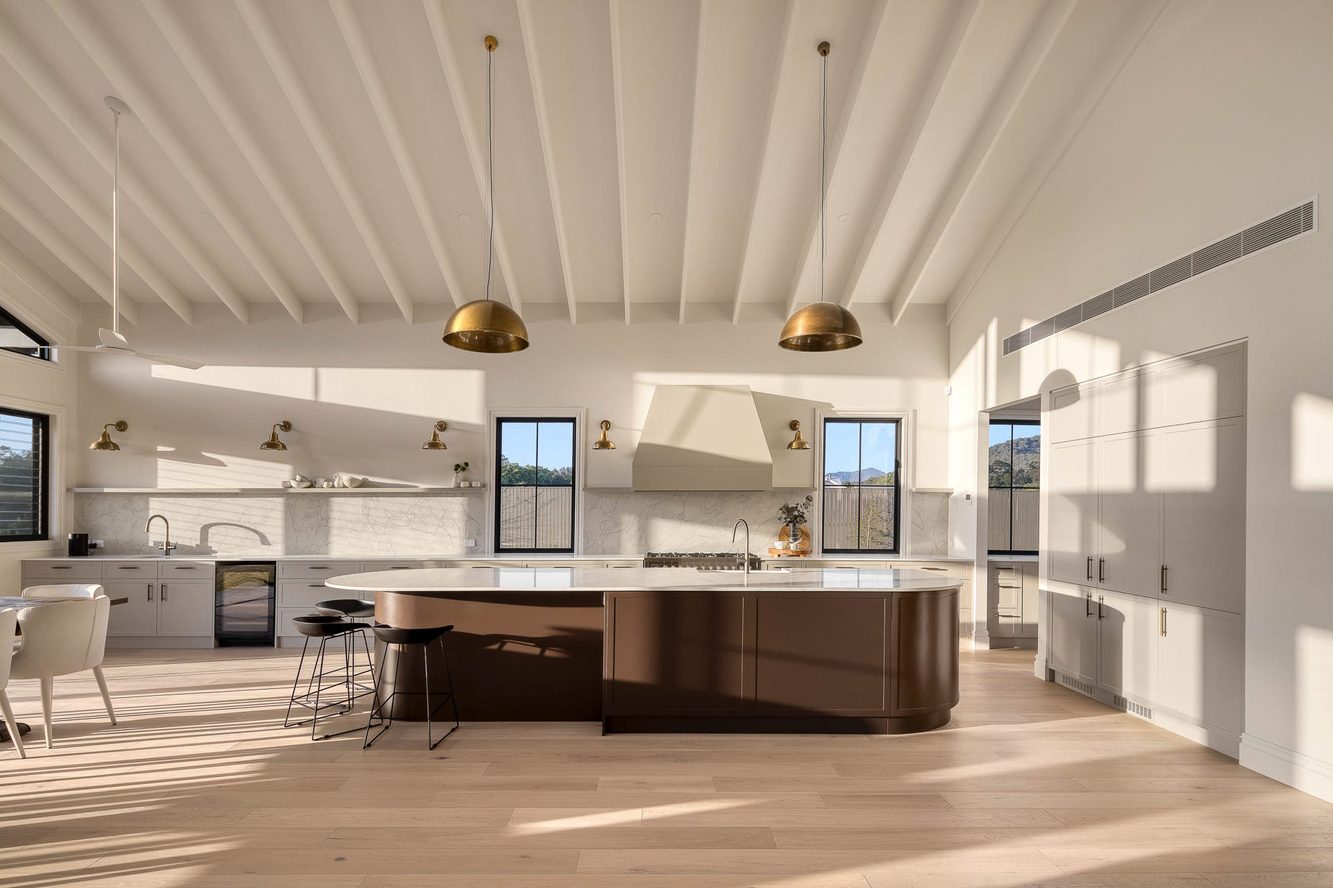 Spacious modern kitchen with large brown island, white countertops, gold pendant lights, and light wooden flooring.