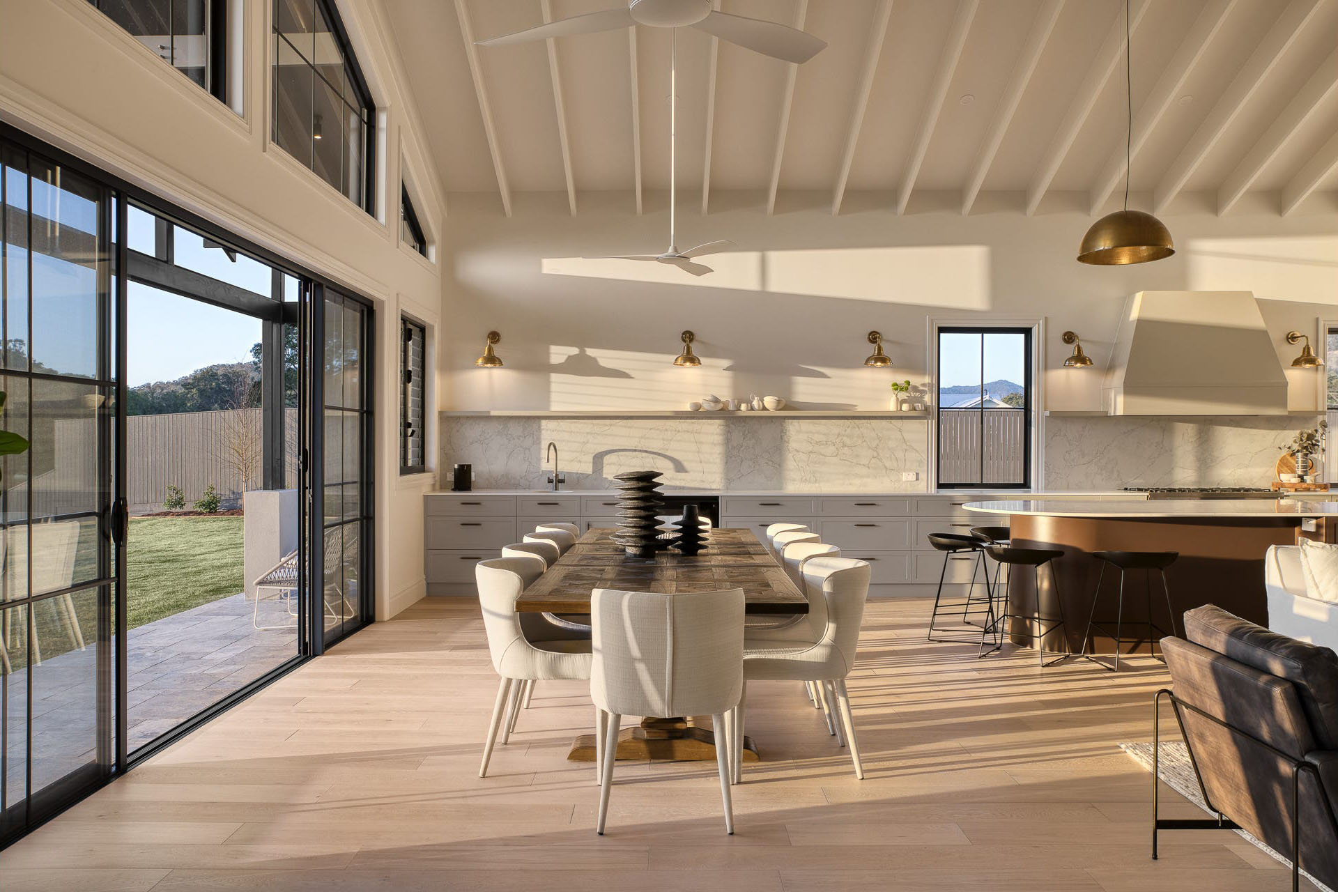 Bright modern dining area with a wooden table, eight beige chairs, large sliding glass doors opening to a patio, and a kitchen with bar stools and brass lighting fixtures.