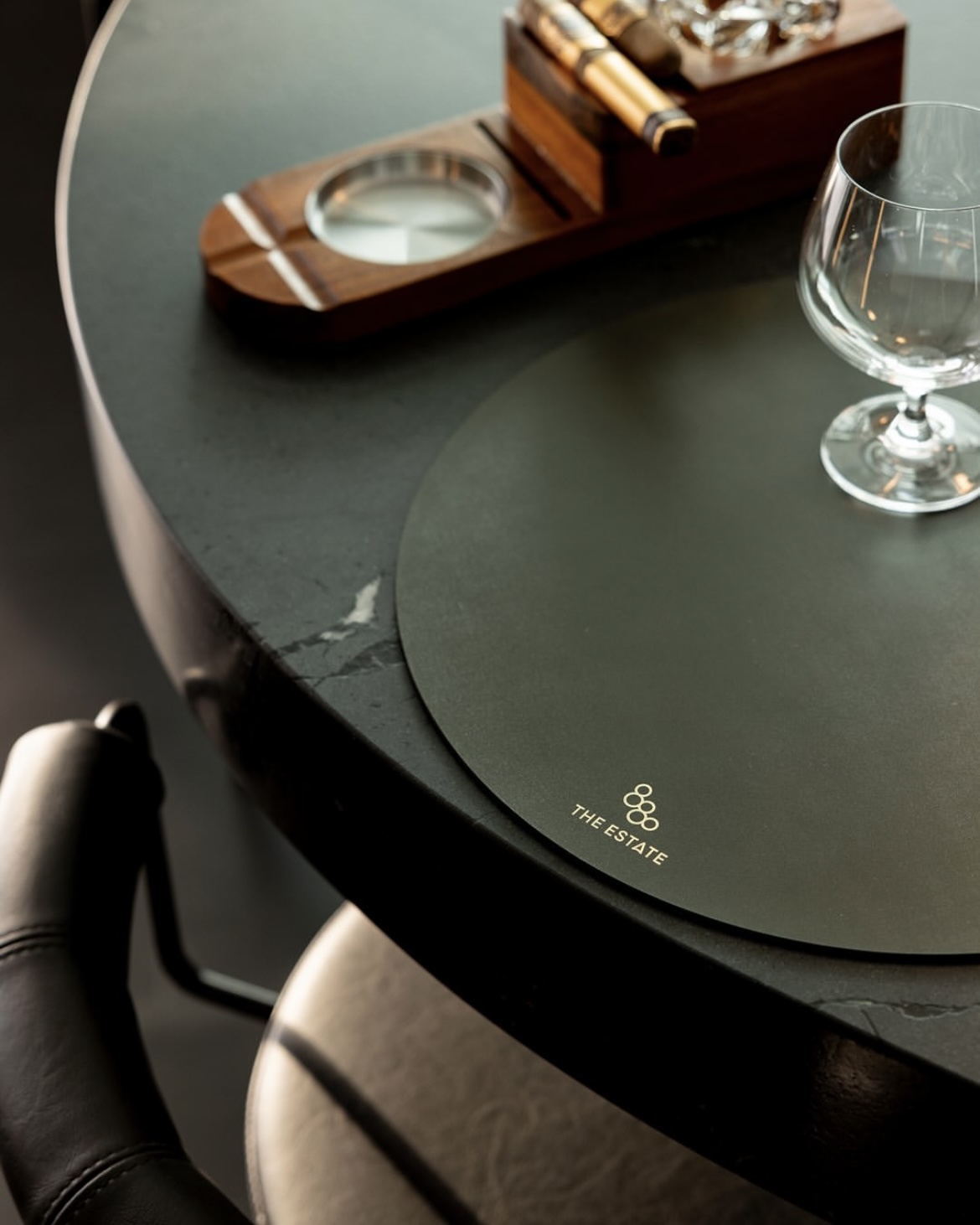Close-up of a dark stone table with a round leather mat labeled 'THE ESTATE', a glass, and a wooden cigar holder.