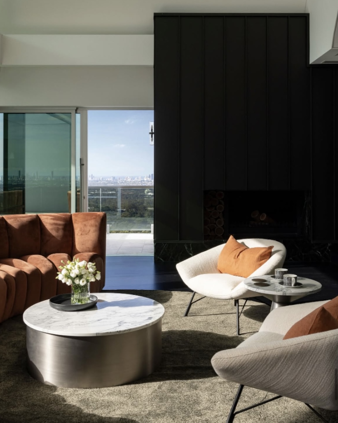 Modern living room with brown velvet sofa, two white armchairs with orange pillows, round marble coffee tables, and a large black fireplace, with a balcony and cityscape view in the background.