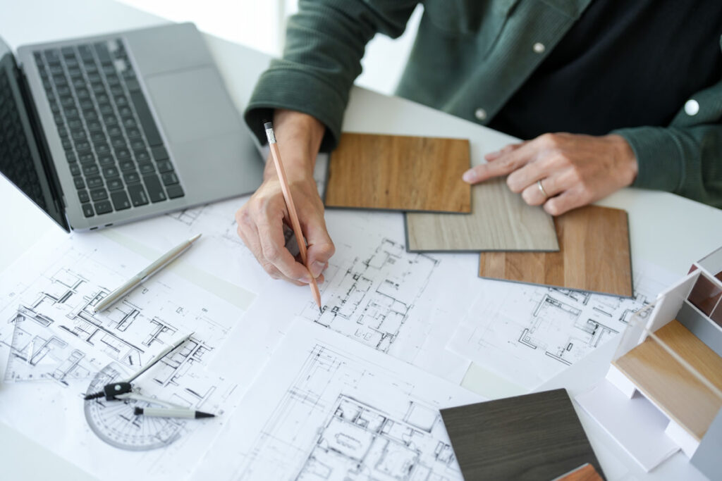 Person working on architectural blueprints with wood samples, laptop, pencil, and drafting tools on a white desk.