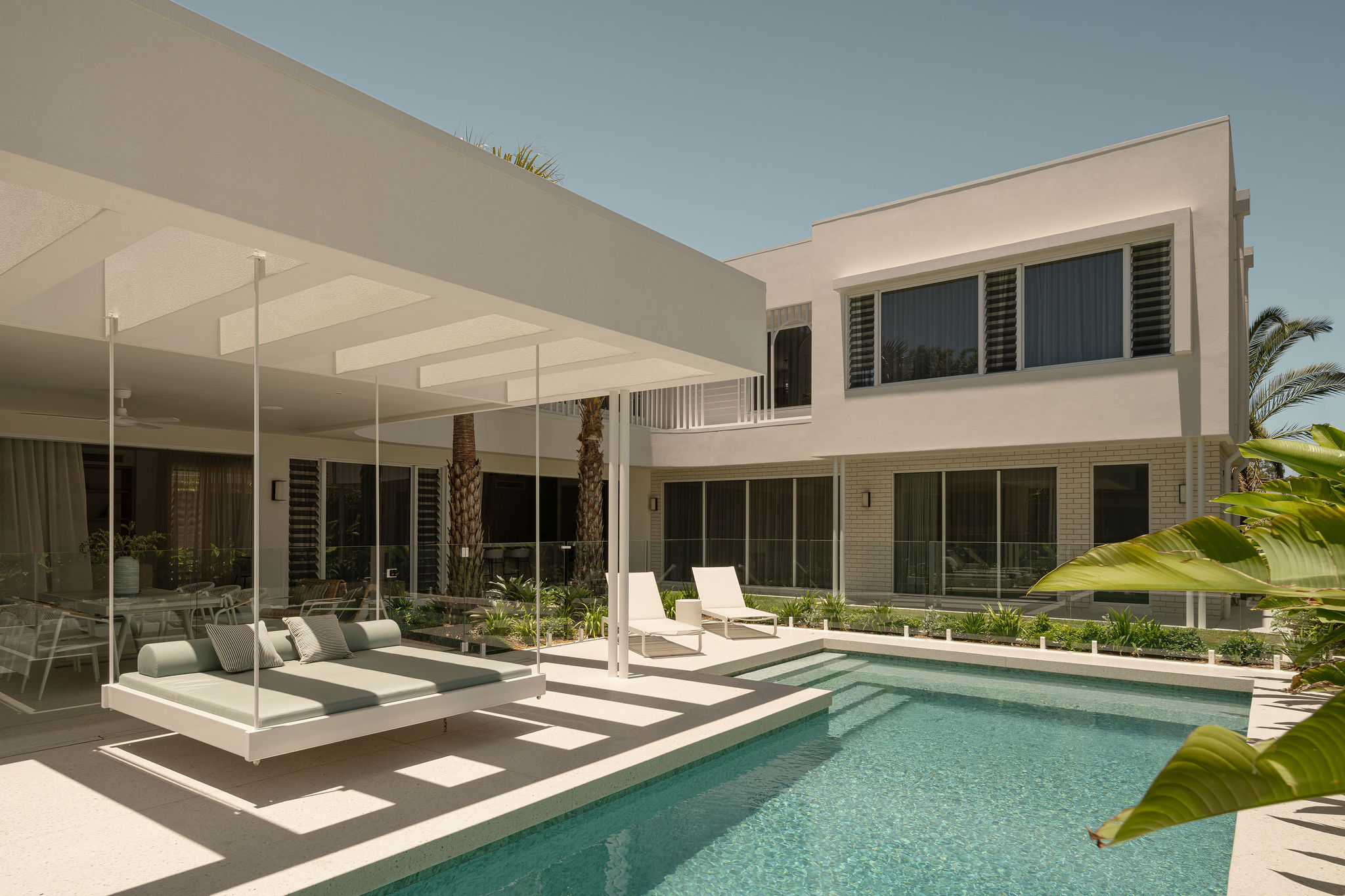 Modern house with a swimming pool, two white lounge chairs, and a suspended outdoor daybed under a shaded pergola.