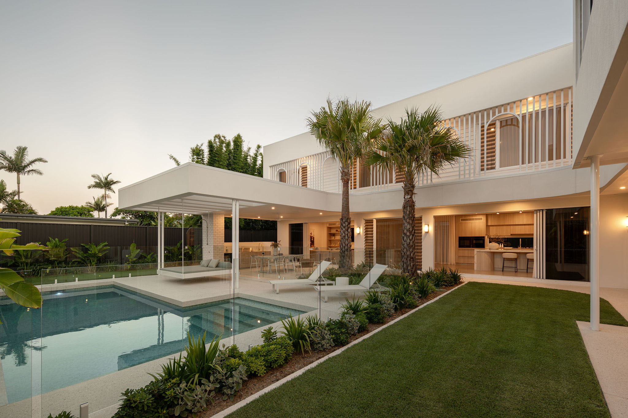 Modern two-story house with large glass sliding doors, outdoor pool, palm trees, and a well-maintained lawn at dusk.