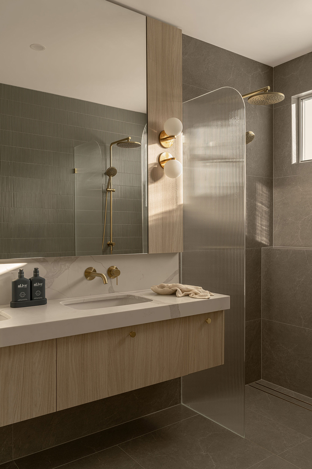 Modern bathroom with beige marble countertop, gold fixtures, wood cabinetry, and a frosted glass shower divider.
