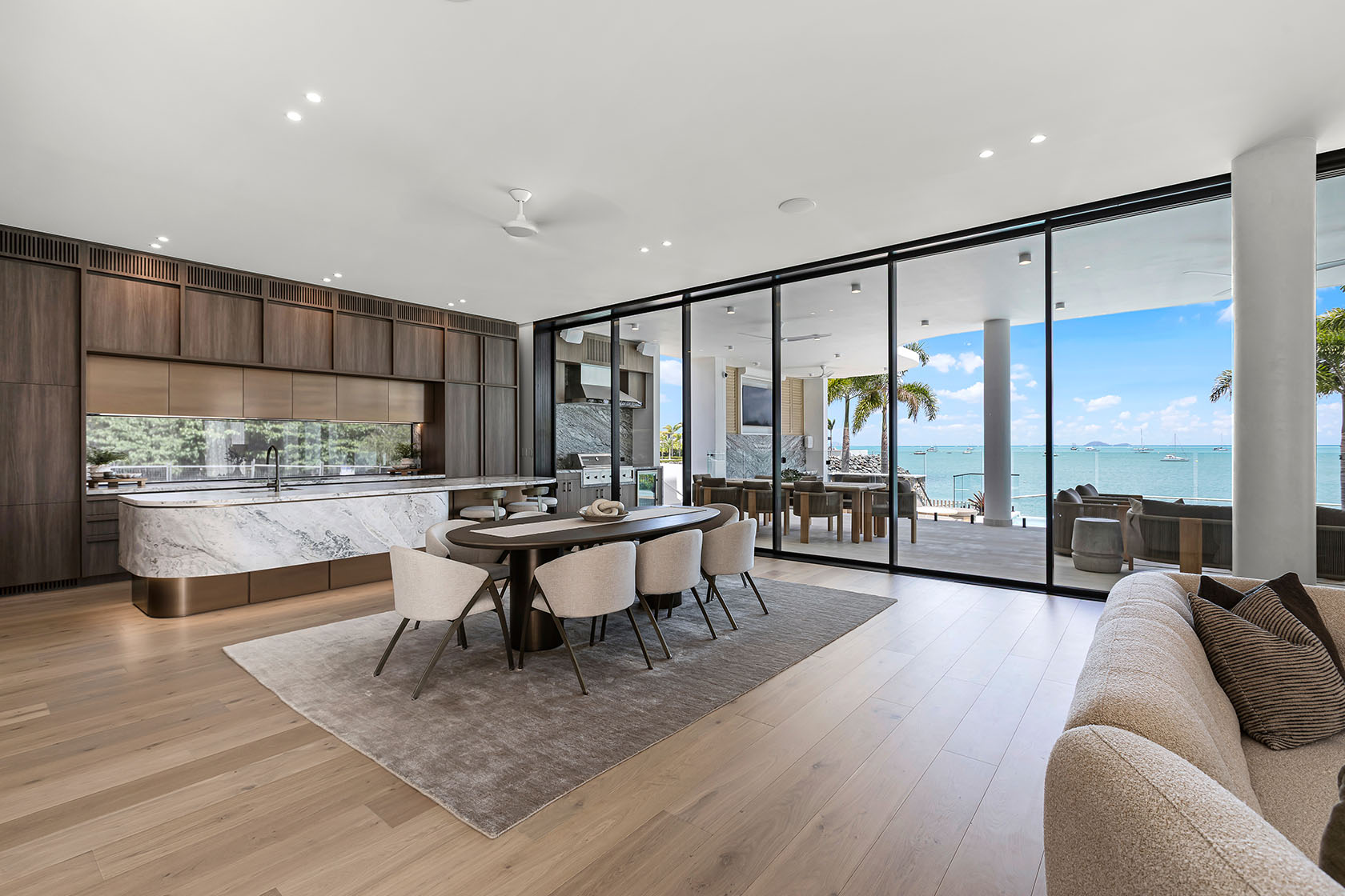 Modern open-plan dining area with beige chairs around dark wooden table, marble kitchen island, and floor-to-ceiling glass doors overlooking a sea view with boats.