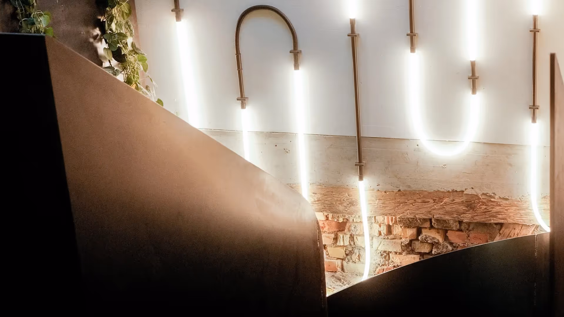 Wand mit rohem sichtbarem Mauerwerk und modernen, kurvigen Leuchten aus Rohren, davor dunkle, gebogene Metallplatten.