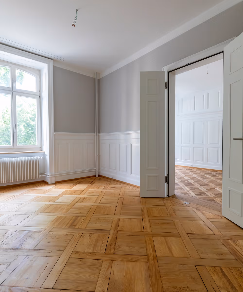 Leerer Raum mit hellem Parkettboden, hohen weißen Wänden mit Paneelen und einem großen Fenster neben einer offenen Tür zu einem ähnlichen Raum.