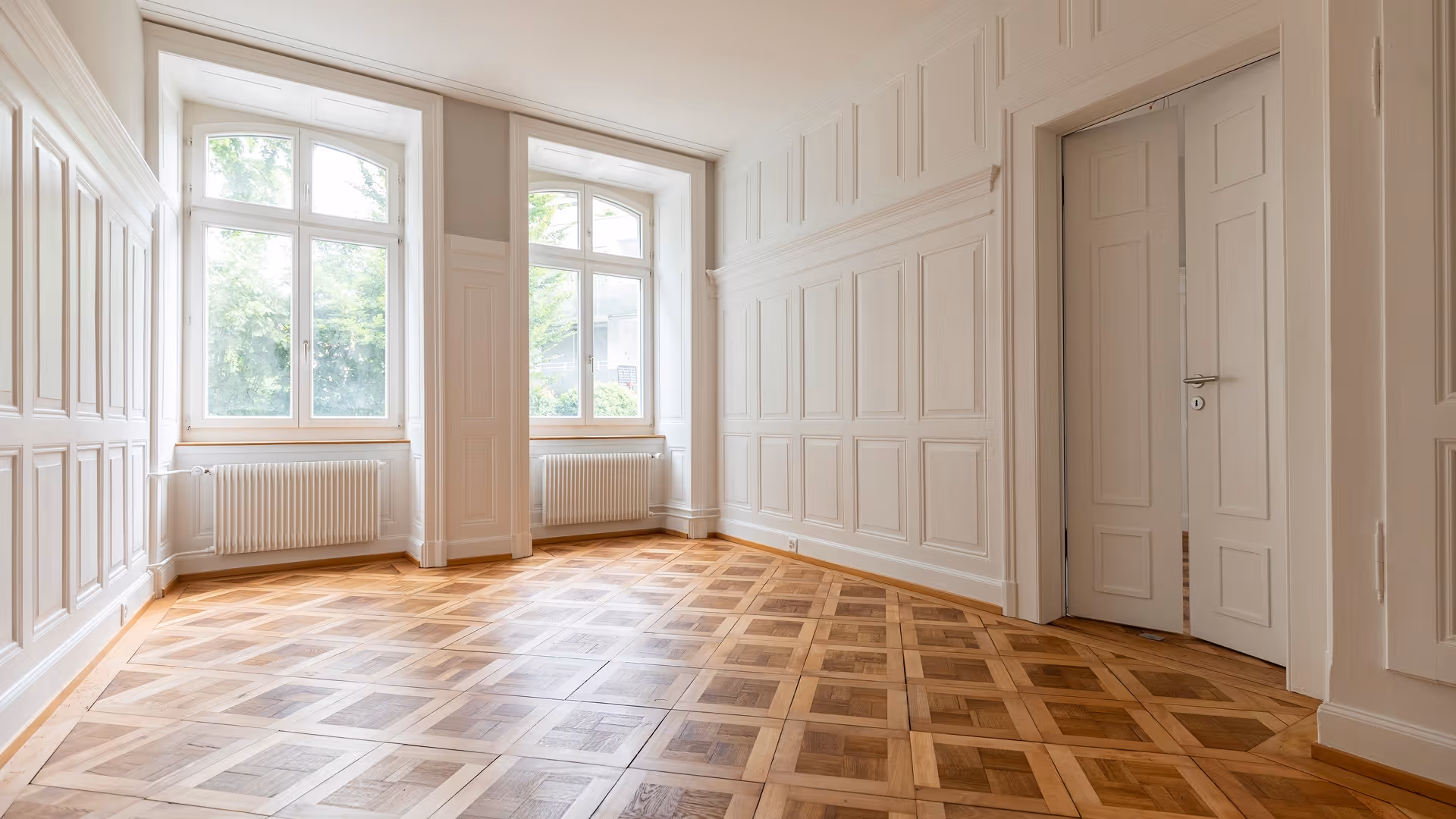 Helles, leeres Zimmer mit großen Fenstern, weißen Wänden mit Zierpanelen und gemustertem Holzparkettboden.