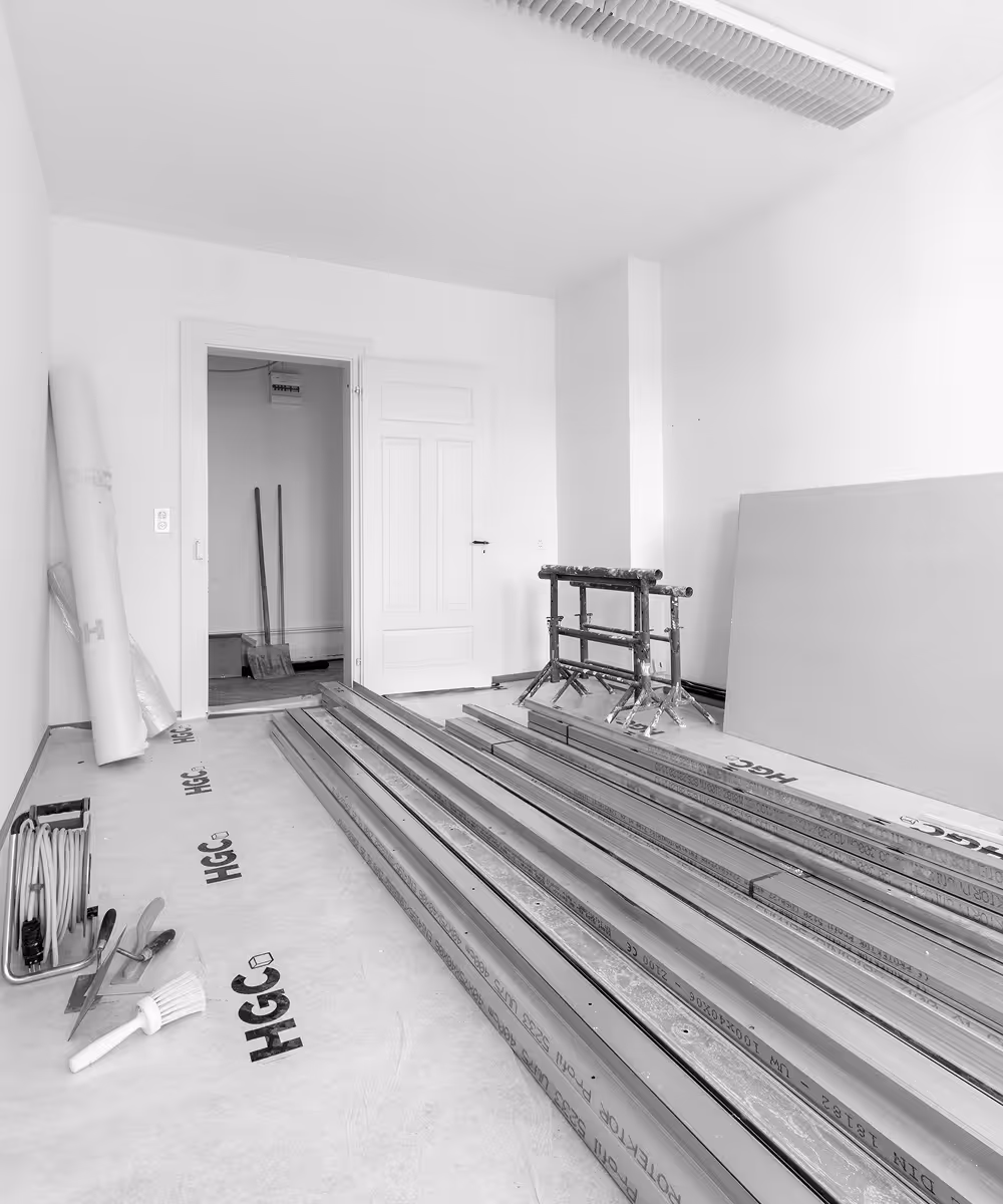 Innenraum mit Baugeräten und Materialien, darunter Holzbalken, Pinsel, Spachtel und gerollte Folie in einem Raum mit weißen Wänden und einer offenen Tür.