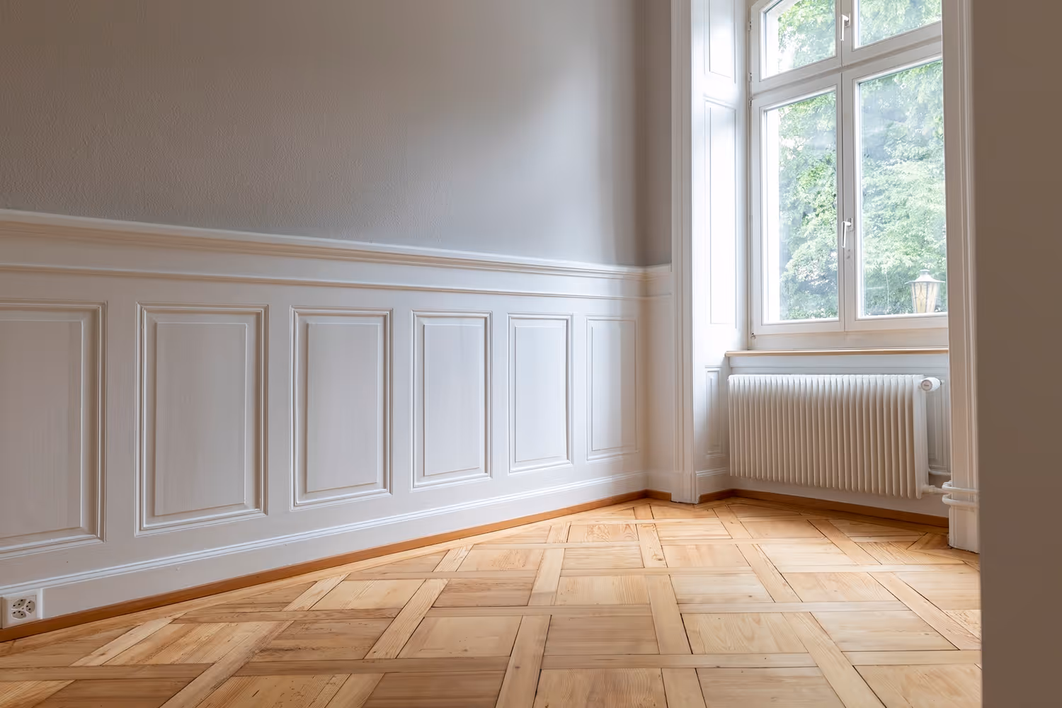 Helles, leeres Zimmer mit weißer Wandvertäfelung, großem Fenster und Parkettboden.