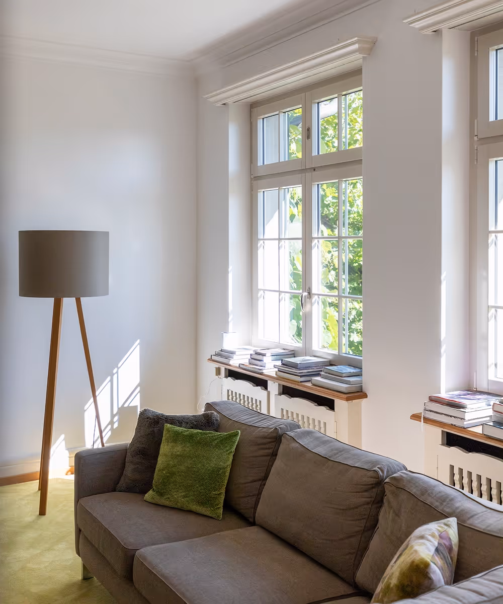 Helles Wohnzimmer mit braunem Sofa, grünen und grauen Kissen, großem Fenster und Stehlampe.