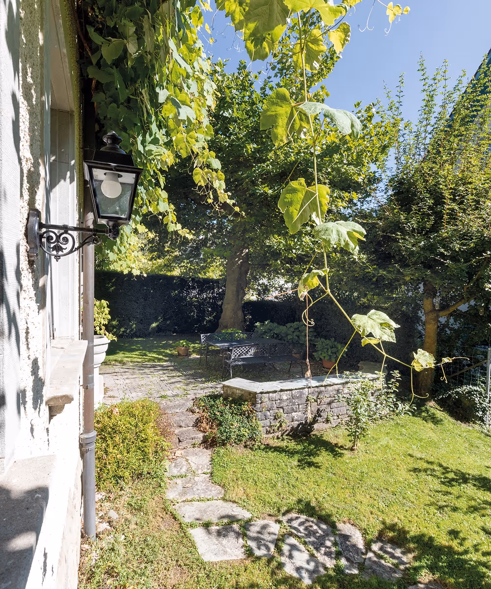 Sonniger Garten mit Steinweg, einer Sitzgruppe aus Metall unter großen Bäumen und einer Hauswand mit einer Außenlaterne.