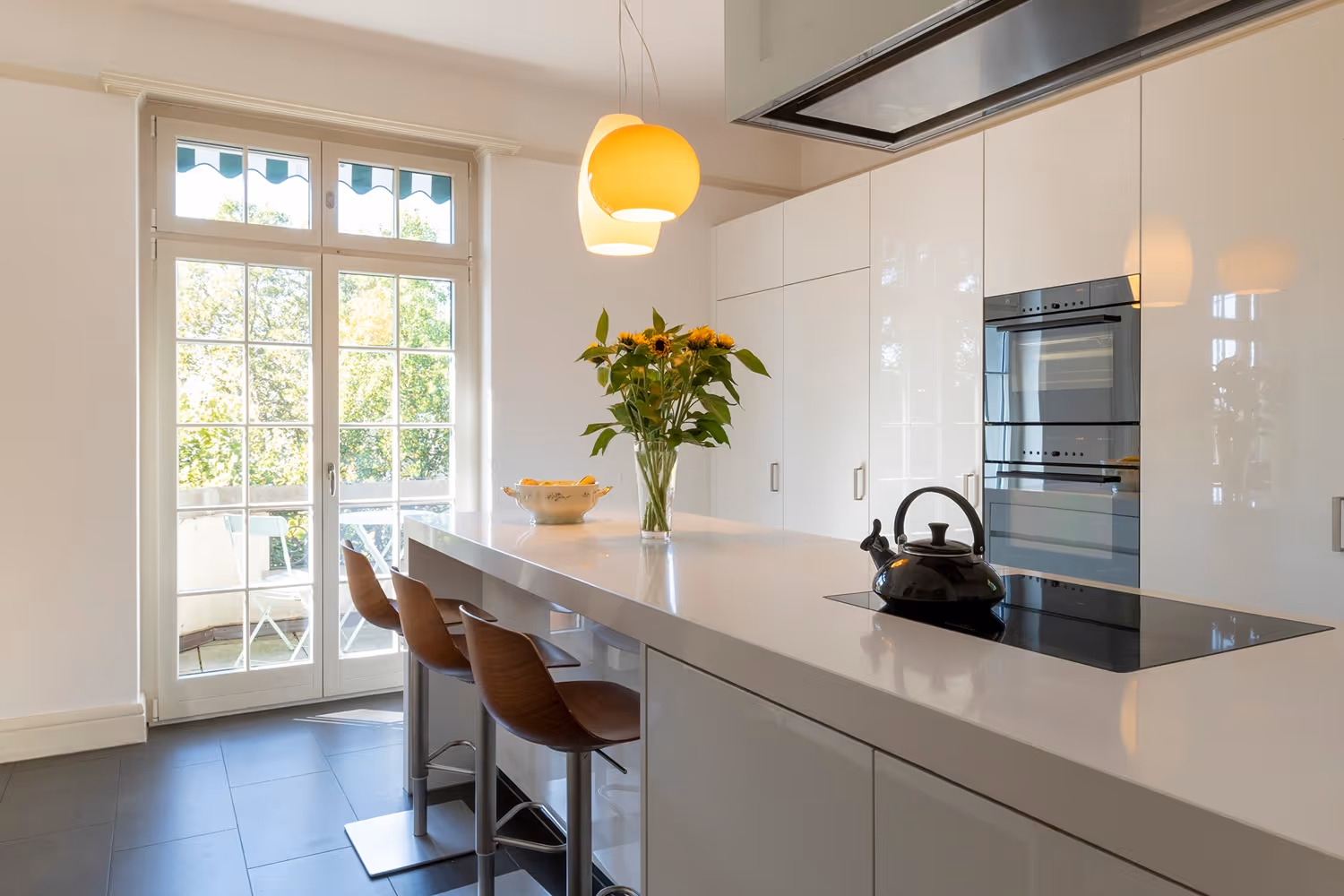 Moderne Küche mit grosser Kochinsel, Kochplatte mit Teekessel, Sonnenblumenstrauss und hellen Holzstühlen vor einer Glasfront mit Blick ins Grüne.