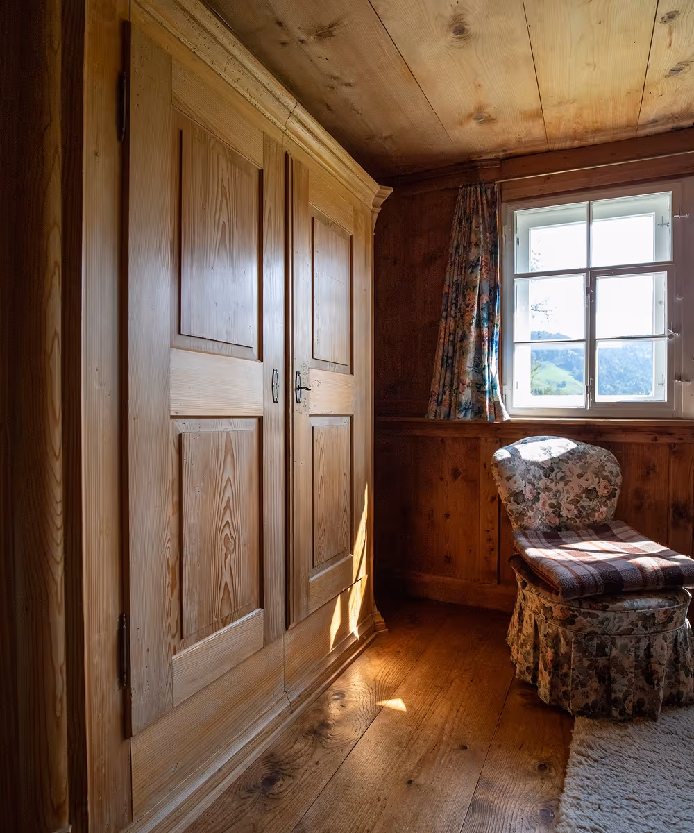 Gemütlicher Raum mit Holzwänden, einem großen Holzschrank, einem blumigen Sessel mit einer karierten Decke und Fenster mit Blick auf eine grüne Landschaft.