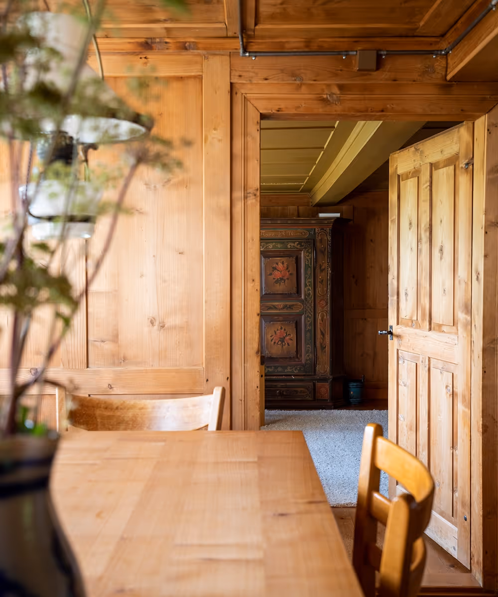 Blick durch eine offene Holztür auf einen Raum mit bemaltem, antik aussehenden Holzschrank, davor ein Holztisch mit Stühlen in einem rustikalen Holzraum.