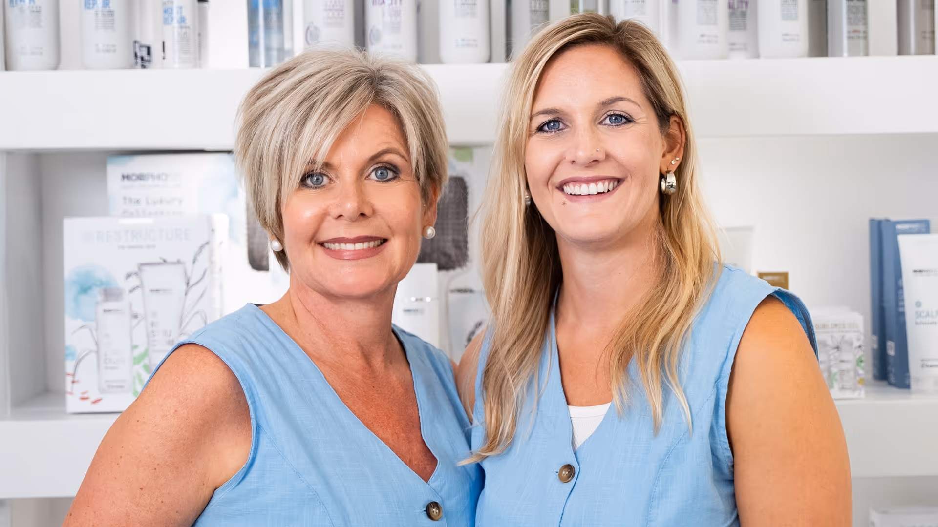 Zwei lächelnde Frauen in hellblauen ärmellosen Blusen vor einem Regal mit Schönheitsprodukten.