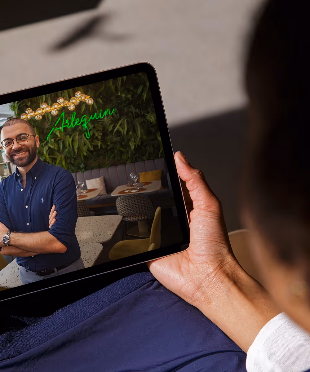 Person hält ein Tablet, auf dem ein lächelnder Mann mit Brille und blauem Hemd vor einer Wand mit Pflanzen und einem 'Arlequin'-Neonschild zu sehen ist.